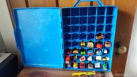 Blue Hot Wheels carrying case open showing several assorted die-cast vehicles in individual slots inside the case.