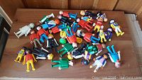 Overview photo showing the full large lot of vintage Geobra Playmobil figures in assorted clothing and colors spread out on a wooden surface.