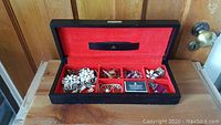 Open black jewelry box showing multiple vintage costume jewelry pieces including rings, brooches, a bracelet, and a necklace organized in compartments with a red velvet interior lining.