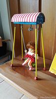 Photo showing the assembled vintage 11-inch swing toy with a doll in red polka dot dress on the swing, metal swing frame with striped canopy.