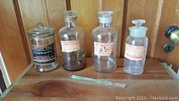Photo showing four vintage glass medical apothecary jars with labels and glass stir stick placed in front, on wooden surface.