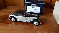 Die-cast model of the Lancia Astura Coupe displayed on a wooden surface with the navy blue tin box in the background with white label.