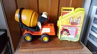 Side and front view of the orange and yellow Fisher-Price cement mixer toy and yellow dollhouse with pink front face designs on a wooden surface.