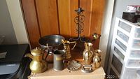 Overview photo showing a collection of antique and vintage metal and brass items including bowl, jugs, vase urns, and a stand arranged against a wooden door.