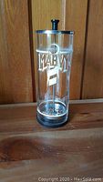 Front view of the clear glass cylindrical container with black knob lid and MARVY logo.