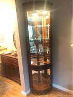 Arc-shaped cherry wood curio cabinet placed against a corner wall. Glass front and sides with interior lighting and five adjustable glass shelves visible. Cherry wood enclosed storage compartment at the bottom.
