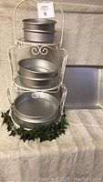 Three-tier white metal wedding cake stand holding 3 round cake pans stacked by size from largest bottom 10 inch to smallest top 6 inch.