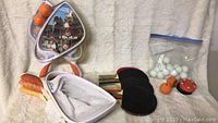 Djubi toss game items including one new pack in a triangular tin with orange tossers, two black-rubber ping pong paddles, and multiple ping pong balls.