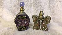 Two metal cast perfume bottles on a white textured fabric background: hexagonal floral bottle with purple stopper and butterfly-shaped bottle with floral rhinestone decoration and clear stopper