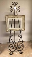 Metal easel style stand with three legs and chain for balance, holding a rectangular distressed white picture frame with carved details, standing against a plain wall and concrete floor.