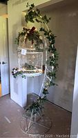 Full view of white metal birdcage hanging on curved metal stand decorated with faux ivy and fairy lights, with artificial flowers and ribbons on birdcage