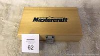 Closed wooden case with Mastercraft logo on lid and metal latch, shown on soft carpet surface with auction tag number 62.
