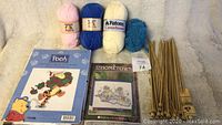 Assortment of knitting needles, balls of wool yarn, and two cross stitch kits photographed together on a soft surface.