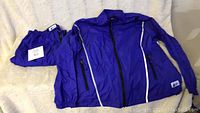 Windbreaker jacket and matching folded wind pants in bright purple color laid out on beige fabric for photo.
