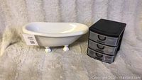 White ceramic clawfoot bathtub decor piece and small black plastic drawer organizer on beige towel background