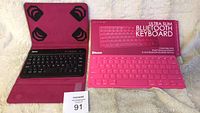 Two small Bluetooth keyboards shown together. Black tzumi keyboard in pink protective stand case on left, pink ultra slim keyboard in original retail box on right.