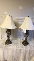 Pair of lamps shown side by side with cream bell-shaped fabric lampshades and ornate metal bases.