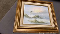Frontal view of vintage framed seascape painting showing lighthouse, sand dunes, and flying seagulls at sunrise or sunset.