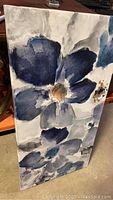 Overall angled view of the large blue floral canvas painting standing vertically showing the flow of blue and gray floral patterns.