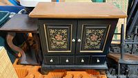 Front view of the black painted cabinet with floral artwork, drawers, white knobs, and natural wood top.
