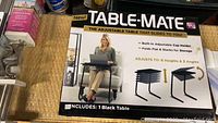 Box showing Table-Mate adjustable table brand, features including 6 heights and 3 angles, built-in cup holder, folds flat for storage, includes 1 black table.
