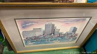 Framed artwork showing a cityscape with buildings and water with sailboats, pastel colors and pen drawing details.
