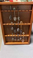 Wooden spoon display rack holding 12 decorative souvenir spoons arranged in three rows.