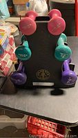 Front view of three pairs of Gold's Gym dumbbells in pink, teal, and purple on display stand.
