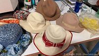 Photo displaying collection of beige, cream, blue, and brown vintage hats and fascinators on a white surface with tags attached to some items.