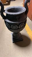 Full side view of the tall black urn showing its shape, pedestal base, and detailed handles.