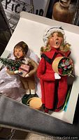 Two Byers Carolers in box, a female with blonde hair in red coat with tambourine, and a boy with brown hair holding Christmas tree and gift box.