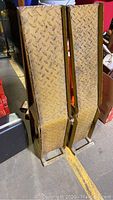 Photo showing pair of upright yellow metal motor vehicle ramps with diamond plate textured surfaces and triangular frame supports.