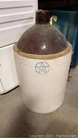 Front view of 5 gallon jug style crock with brown glazed top and blue star symbol on beige body.