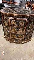 Front view of black painted small cabinet with detailed floral decorations on drawers and cabinet doors.