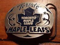 Front view of the oval pewter belt buckle showing the Toronto Maple Leafs logo in blue and silver with decorative stars, with 'Toronto' scripted above and 'Maple Leafs' at the bottom.