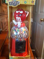 Front view of M&M's red character candy dispenser and coin bank in original packaging, showing the clear candy globe filled with colorful plastic candies and the red character figure on top.