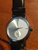 Close-up of one watch showing round silver-tone case, two-tone dial with small sub-dial, and black leather strap.