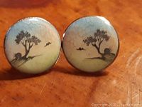 Close-up of one hand painted enamel earring showing tree and birds scene with crackled enamel surface.