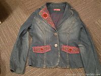 Full view of the ladies denim jacket laid flat showing front side with embroidery on collar and pocket flaps, metal buttons, fitted silhouette