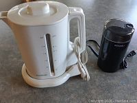 White Proctor Silex electric kettle with water level markings and power cord alongside black Regal coffee/spice grinder on countertop.