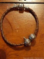 Full view of the braided leather bracelet showing the silver clasp with 'PAND' engraving and two charms attached.