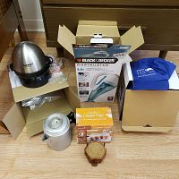 Overview photo of all items in the lot including Black & Decker Dustbuster, Copper Chef Copper Eggs box, Cuisinart egg cooker, metal coffee pot, and wooden trivet on the floor.