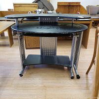 Front view of black computer desk with curved top and multiple shelves, metal perforated side panels, and caster wheels.