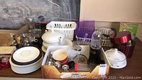 Wide view of kitchen items including plates, glasses, condiment shakers, covered casseroles, soup tureen and Magic Bullet blender base with cups and lids grouped on table.
