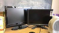 Photo showing Acer and Samsung monitors side by side on a surface, power cables visible in front of Acer monitor, fan on right.