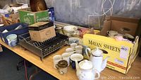 Overview of the table showing ceramic mugs, metal dish rack, teapots, and boxed items