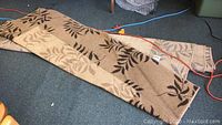 Photo showing folded beige rug with brown and cream leaf pattern on blue carpeted floor with orange extension cord.