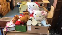 Several plush toys including two large Hello Kitty dolls, a Doctor Seuss plush, and a Hello Kitty lunchbox in a cardboard box on a surface.