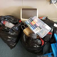 Two large black plastic bags filled with yarn and a beige waste basket behind; a clear plastic box of polyester thread spools displayed on one bag.
