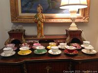 Wide view of 13 assorted fine china cup and saucer sets displayed on a wooden sideboard.